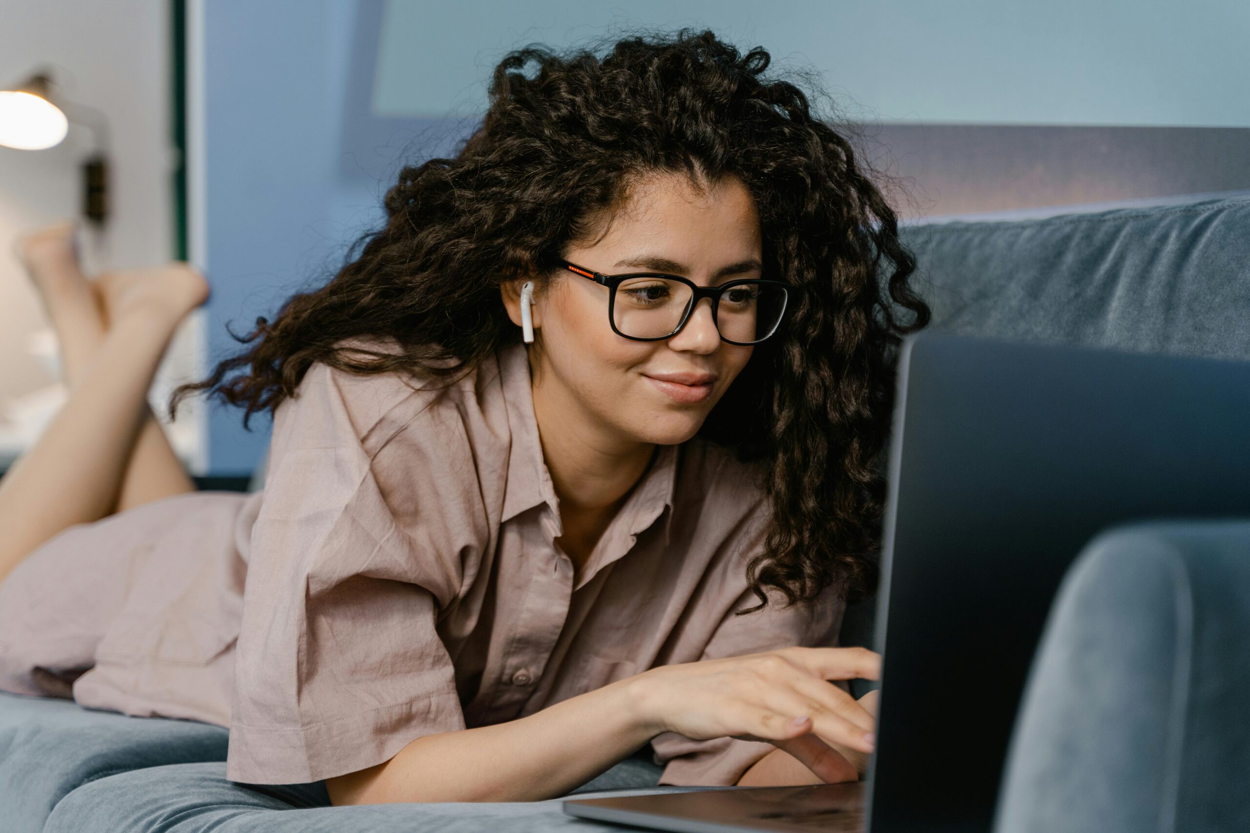 Smiling woman with curly hair and eyeglasses working on a laptop indoors.